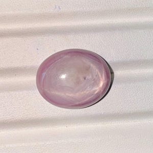 May include: A polished, oval-shaped gemstone with a soft, translucent appearance. The gem features a blend of pale pink and white hues, with subtle internal patterns. The stone is set against a white, textured surface.