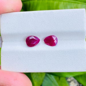 May include: Two pear-shaped, faceted, deep red gemstones. The gems are displayed on a white surface, with a green background. The gems are a vibrant shade of red, with visible facets that catch the light.