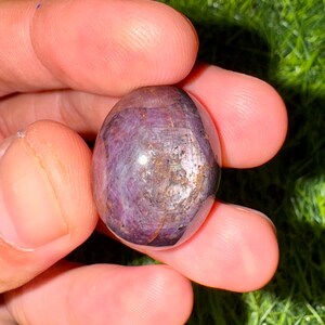 May include: A smooth, oval gemstone in shades of deep purple, held between fingers. The stone displays a play of light and colour variations, with a metallic sheen on its surface. The rounded form of the gemstone is highlighted, showcasing its natural beauty.