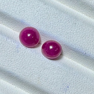 May include: Two round, polished, deep magenta gemstones rest on a textured white surface. The gems have a glossy finish, reflecting light. The background is a white, textured material.