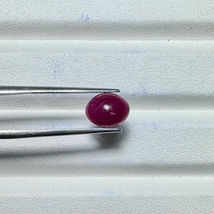 May include: A polished, oval-shaped, deep red gemstone is held by a pair of silver tweezers. The gem has a smooth, reflective surface and is set against a white, textured background. The tweezers are positioned to hold the gem securely.
