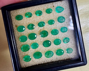 May include: A collection of emerald gemstones in various oval and pear shapes, displayed in a black framed case. The gemstones are a vibrant green color and are set against a light-colored foam backing.