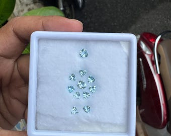 May include: A white plastic box containing several light blue, heart-shaped gemstones. The gemstones are arranged on a white surface. The box is held in a person's hand, with a blurred background of a scooter and other objects.