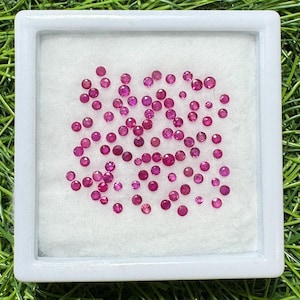 Puede incluir: Una colección de pequeñas gemas redondas de color rojo intenso, posiblemente rubíes, se exhiben dentro de un contenedor cuadrado blanco con interior blanco. Las gemas varían en tamaño y están dispersas sobre la superficie blanca. El fondo es verde.