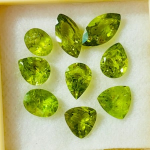 Op de afbeelding: Een verzameling van negen gefacetteerde, lichtgroene peridot edelstenen in verschillende vormen, waaronder ovaal, peervormig en marquise. De edelstenen worden gepresenteerd in een crèmekleurige doos, die hun helderheid en levendige kleur benadrukt.
