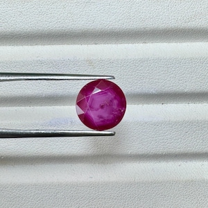 May include: A faceted, round, deep red gemstone held by silver tweezers. The gem has a brilliant cut, reflecting light across its many facets. The stone's color is a rich, saturated red, with a slight pink hue. The tweezers are positioned against a white background.