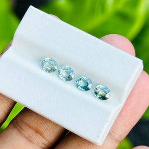 Puede incluir: Cuatro piedras preciosas ovaladas en tonos de verde y azul se muestran sobre una superficie blanca. Las piedras preciosas están dispuestas en una fila de izquierda a derecha, con la piedra preciosa más grande a la izquierda y la más pequeña a la derecha.
