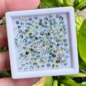 May include: A collection of small, round gemstones in shades of blue, green, and yellow are displayed in a white square container. The gemstones are of varying sizes and are scattered across the white surface, creating a sparkling effect.