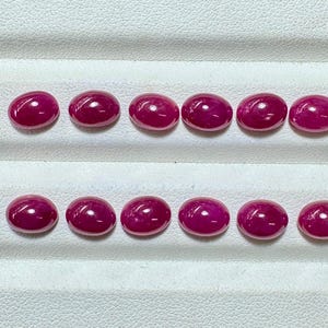 Può includere: Una collezione di dodici gemme ovali e lucide. Le gemme sono di un colore rosso intenso e vibrante, con una superficie liscia e riflettente. Sono disposte in due file parallele su una superficie bianca e strutturata. Le gemme sembrano essere rubini.