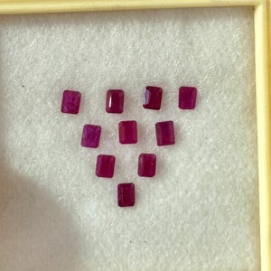 May include: A collection of ten rectangular, deep red gemstones arranged in a display case. The gems are set against a white background within a light yellow frame. The stones have a polished, reflective surface.