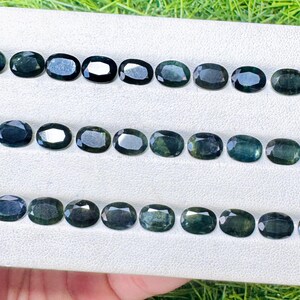 May include: An array of oval-cut, dark teal gemstones arranged on a white surface. The gems are displayed in three neat rows, each stone exhibiting a polished, reflective surface. The overall presentation suggests a collection of precious stones, possibly for jewelry making.