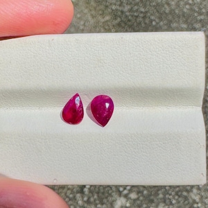 May include: Two vibrant, pear-shaped, deep red gemstones displayed on a white textured surface. The gems are positioned side-by-side, showcasing their polished facets and rich color. The background is a neutral gray.