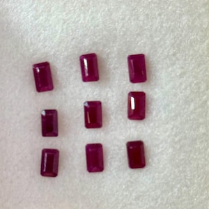 May include: Nine rectangular, deep red gemstones are arranged on a white surface. The gems have a polished, reflective surface, and are evenly spaced. The color is a rich, saturated red, suggesting they are rubies or similar precious stones.