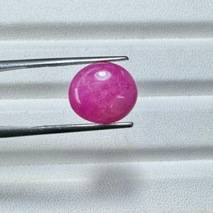 May include: A round, polished, hot pink gemstone is held by a pair of silver tweezers. The gemstone has a smooth, glossy surface and is set against a white background. The tweezers are positioned to hold the stone securely.
