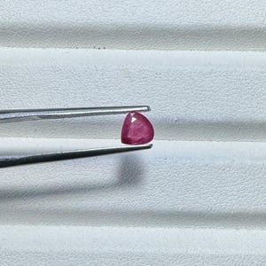 May include: A faceted, triangular-shaped, deep red gemstone is held by a pair of silver tweezers. The gem is positioned against a white, textured background, showcasing its vibrant color and cut. The tweezers are in sharp focus.