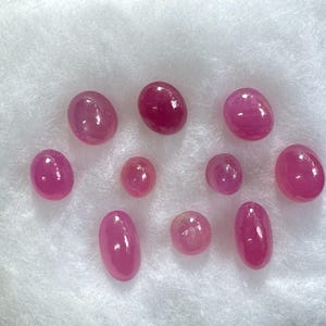 Peut inclure: Une collection de pierres précieuses roses polies, de forme ovale et de tailles variées. Les gemmes sont présentées sur une surface blanche et duveteuse, mettant en valeur leurs surfaces lisses et réfléchissantes et leur couleur riche.