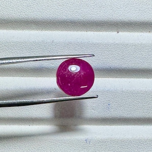 May include: A vibrant, round, ruby gemstone held by silver tweezers. The gemstone is a deep, saturated pink-red color, with a smooth, polished surface. The tweezers are positioned against a textured white background.