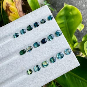 May include: A collection of oval-cut gemstones arranged in three rows on a white display board. The stones exhibit various shades of blue, teal, and green, featuring faceted surfaces that reflect light. Each gemstone is distinct in color and size.
