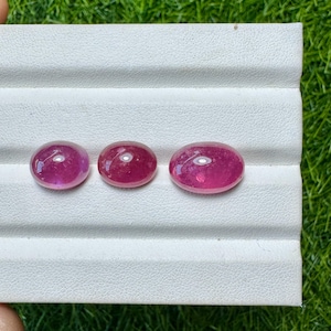 May include: Three oval-shaped, polished gemstones in varying sizes. The gems are a deep, vibrant pink-red color, with a smooth, reflective surface. They are displayed on a white, textured surface against a backdrop of green grass.