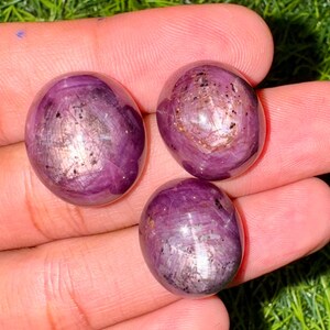 May include: Three polished, oval gemstones in a deep purple colour. Each stone has a shimmering, iridescent quality with internal patterns. The gems are displayed against a background of green grass.