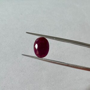 May include: A faceted, oval-shaped, deep red ruby gemstone is held by a pair of silver tweezers against a white background. The ruby's polished surfaces reflect light, showcasing its rich color and clarity. The tweezers provide a close-up view of the gem.