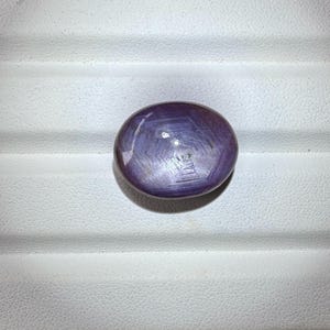 Könnte beinhalten: Ovalförmiger, blau-violetter Edelstein mit Sterneneffekt. Die polierte Oberfläche reflektiert das Licht und erzeugt ein sechszackiges Sternmuster. Der Edelstein liegt auf einem weißen, strukturierten Hintergrund.