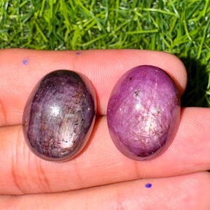 May include: Two oval-shaped, polished gemstones. One is a deep purple with reflective streaks, the other a vibrant pinkish-purple. The stones are smooth and appear to be held in a hand, with green grass in the background.