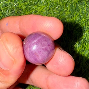 Op de afbeelding: Een gepolijste, bolvormige edelsteen in roze en paarse tinten, vastgehouden tussen vingers. De steen heeft een glad oppervlak met subtiele interne patronen en een reflecterende glans, wat suggereert dat het een natuurlijk kristal of mineraal is.