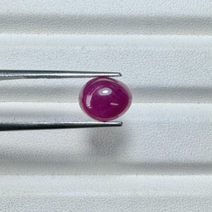 May include: A polished, oval-shaped ruby gemstone, held by tweezers. The ruby is a deep, saturated red color with a smooth, reflective surface. The tweezers are silver-colored and hold the gemstone against a white background.