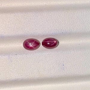 Puede incluir: Dos gemas ovaladas de color rojo intenso con efecto de estrella, sobre fondo blanco. Probablemente rubíes, a menudo utilizados en joyería. Cada gema mide aproximadamente 1 cm de largo.