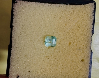 May include: A small, square-cut, pale blue gemstone is presented on a yellow foam pad inside a black container. The gemstone has a faceted surface, reflecting light. The image is a close-up, highlighting the gem's clarity and colour.