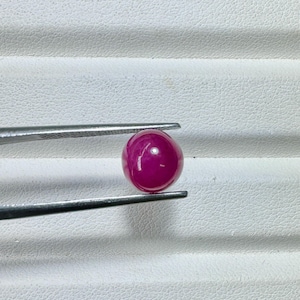 May include: A polished, round, deep red gemstone, held by silver tweezers. The gem has a smooth, reflective surface, and is set against a white, textured background. The gemstone is a vibrant, eye-catching piece.