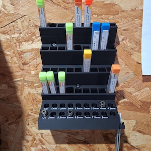 May include: A black metal drill bit organizer with multiple compartments and labeled holes for various sizes. It holds several drill bits with colorful tops, including green, orange, blue, and yellow. The organizer is mounted on a wooden surface.