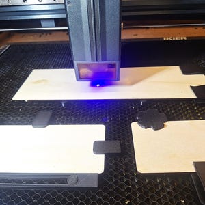 May include: A laser cutter in operation, with a blue laser beam focused on a light-coloured wooden surface. The machine has a dark grey housing and is positioned above the wood. Several black fixtures are holding the wood in place.