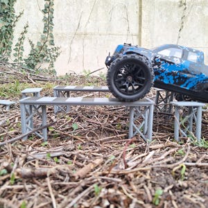 Könnte beinhalten: Ein blau-schwarzer ferngesteuerter Monstertruck steht auf einer Reihe grauer Miniaturplattformen. Die Plattformen befinden sich in einer natürlichen Umgebung mit Zweigen und Blättern.