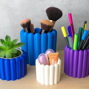 Puede incluir: Una colección de recipientes coloridos y acanalados que contienen varios artículos. Un recipiente azul contiene brochas de maquillaje, un recipiente blanco contiene cosméticos y un recipiente morado contiene bolígrafos y rotuladores. Un recipiente azul contiene una planta suculenta. Los recipientes están sobre una superficie de madera.