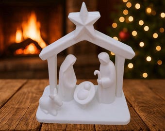 Puede incluir: Un belén de cerámica blanca con María, José y el niño Jesús bajo un refugio coronado por una estrella. Un burro está situado a la izquierda. La escena está ambientada sobre una superficie de madera con una chimenea y un árbol de Navidad al fondo.