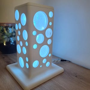 May include: A white, rectangular lamp with a square base. The lamp features various sized circular cutouts that emit a soft blue light. The lamp is plugged into an electrical outlet. The lamp is sitting on a wooden surface.