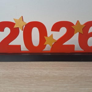Peut inclure: Décoration en bois rouge avec l'année 2026 et des étoiles dorées. Les chiffres sont en gras et montés sur une base noire. Les étoiles sont placées entre les chiffres. Idéal pour un événement du Nouvel An ou une célébration.