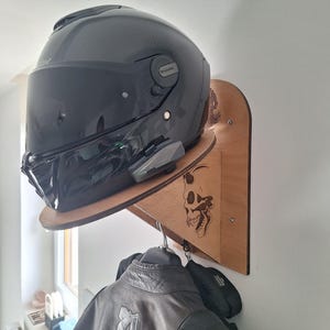 May include: A black motorcycle helmet with a tinted visor rests on a wooden shelf attached to a wall. Below, a black leather jacket hangs from a hook. The shelf features a skull design.