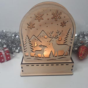 May include: A wooden Christmas lantern with a winter scene cut out. The design includes deer, trees, mountains, and snowflakes. The lantern is lit from within, creating a warm glow. A small gift box and a red ornament are in the background.