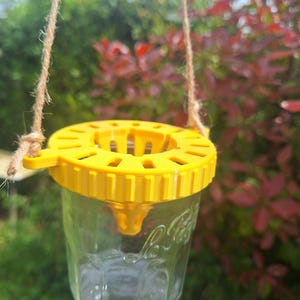 Op de afbeelding: Een heldere glazen pot met een gele plastic deksel en een touw om op te hangen. De deksel heeft een reeks gaten en een kleine tuit. De pot hangt tegen een achtergrond van groen en rood gebladerte.
