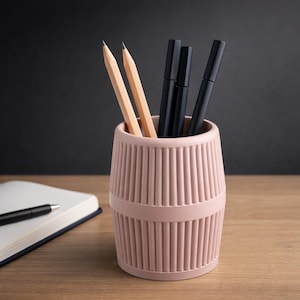 Peut inclure: Un pot à crayons rose avec des rainures verticales contient deux crayons et trois stylos noirs. Un cahier ouvert avec un stylo repose sur une surface en bois, sur fond sombre.
