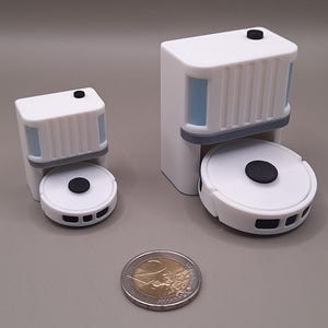 Puede incluir: Dos modelos en miniatura de robots aspiradores blancos y grises con detalles negros. Los modelos tienen una parte superior rectangular y una base circular. Una moneda de dos euros está al lado de los modelos para comparar el tamaño.