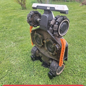 May include: A robotic lawn mower positioned vertically on its side, revealing the underside with two circular cutting blades, four omnidirectional wheels with black treads, a white top handle, and orange side support arms on a green lawn.