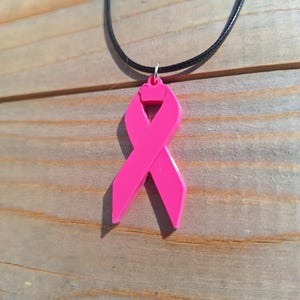 Peut inclure: Un collier pendentif ruban rose vif. Le pendentif, symbole de sensibilisation au cancer du sein, est suspendu à un cordon noir avec un fermoir argenté. Le collier est présenté sur un fond de planches de bois.