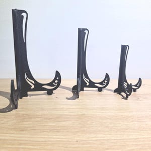 Puede incluir: Tres expositores negros de diferentes tamaños están dispuestos sobre una superficie de madera clara. Cada soporte tiene un diseño decorativo y curvo en la base y una parte trasera alta y delgada. Los soportes están diseñados para sostener platos u otros objetos.