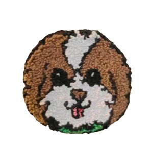 May include: A round, textured dog face design in brown, white, black, and pink. The dog has a white patch on its forehead, black eyes, and a pink tongue. The design is set against a white background.