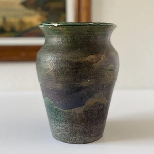 May include: A handcrafted, small ceramic vase with a textured surface. The vase features a color palette of dark green, brown, and purple hues. The vase has a slightly flared rim and a rounded body, suitable for displaying flowers or as a decorative piece.