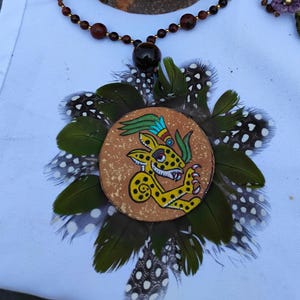 Può includere: Una collana artigianale con un ciondolo circolare raffigurante un giaguaro giallo, circondato da piume verdi e macchiate. La collana ha una catena di perline con accenti marroni e dorati, che mostra uno stile artistico unico.
